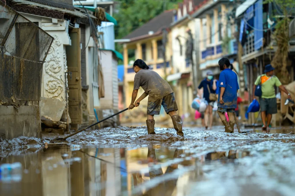 People in boots clear thick mud and floodwater from a street lined with buildings.