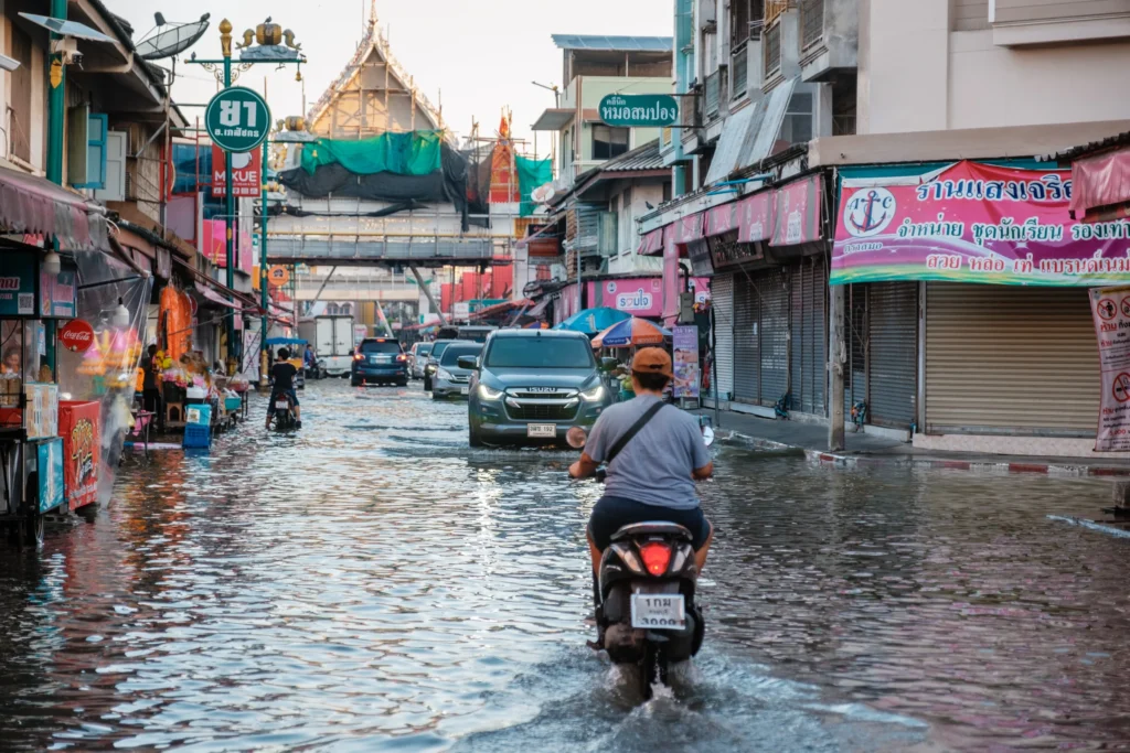 A motorcyclist and cars travel through floodwater on a Thai market street lined with small shops.